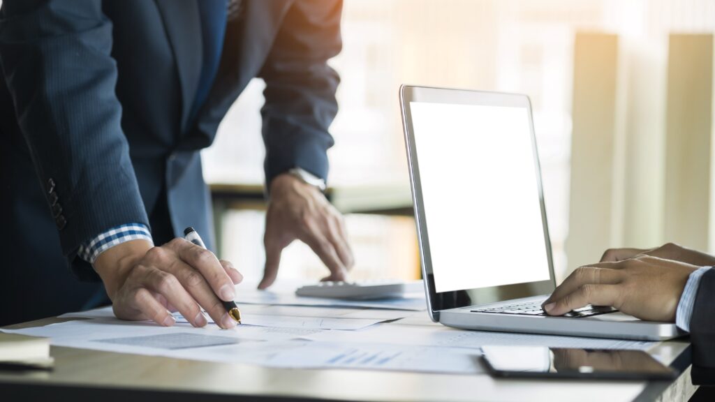 Business professional leaning over desk, writing something, someone else typing on laptop